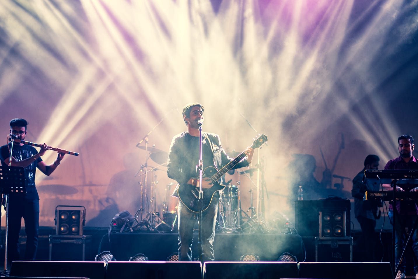 A full band on stage, silhouetted in a wash of purple light and haze.